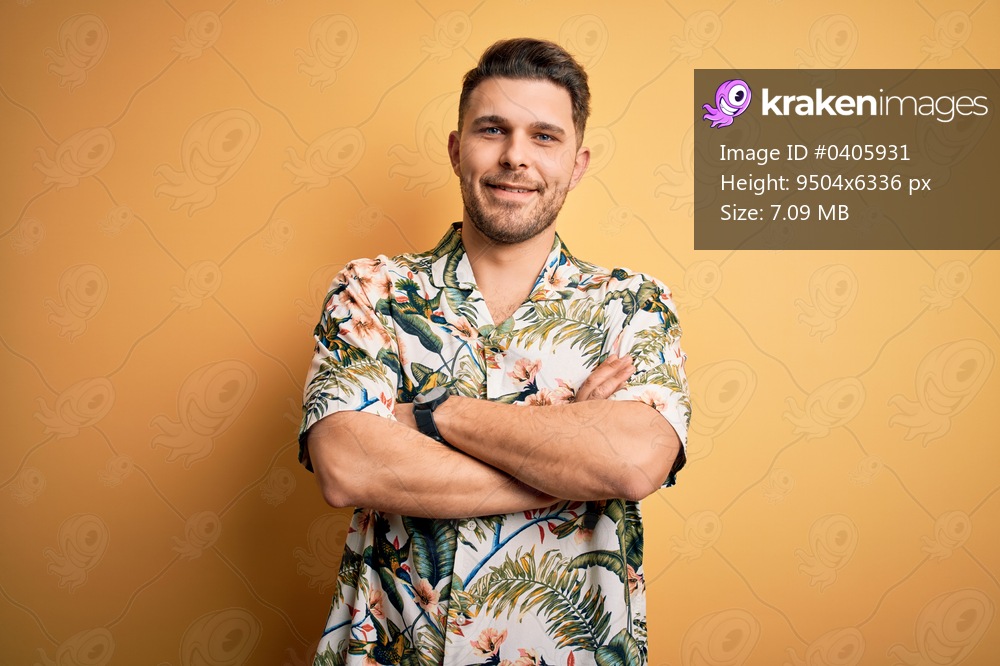 Young man with blue eyes on vacation wearing floral summer shirt over yellow background happy face smiling with crossed arms looking at the camera. Positive person.