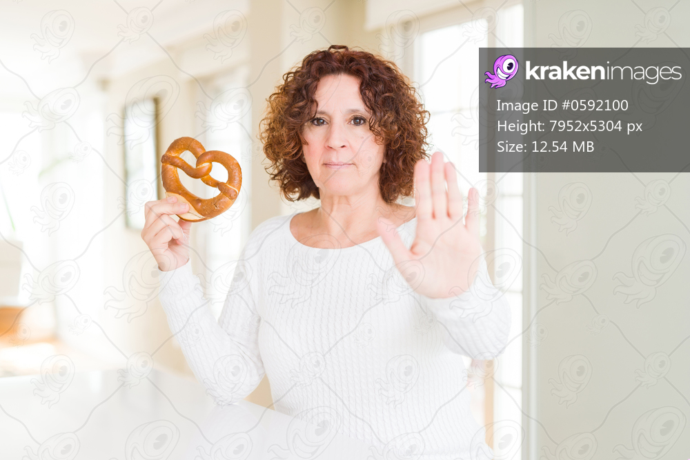 Senior woman eating german salty pastry pretzel for octoberfest with open hand doing stop sign with serious and confident expression, defense gesture