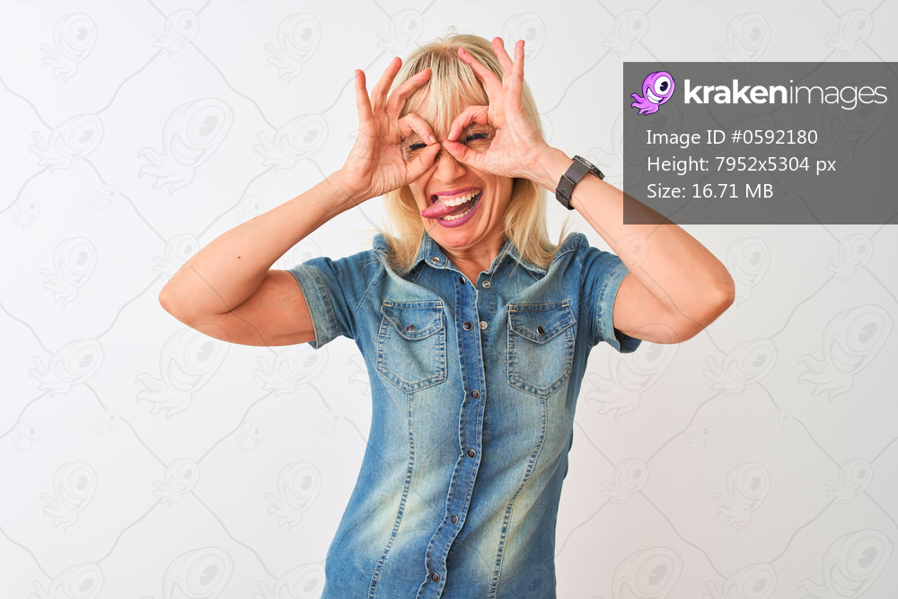 Middle age woman wearing casual denim shirt standing over isolated white background doing ok gesture like binoculars sticking tongue out, eyes looking through fingers. Crazy expression.