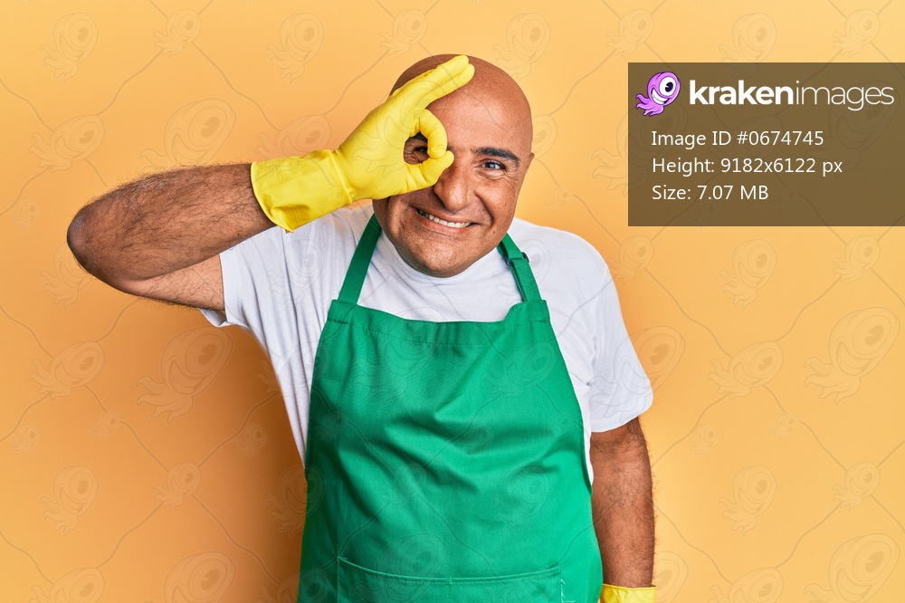 Mature middle east man wearing cleaner apron and gloves smiling happy doing ok sign with hand on eye looking through fingers 