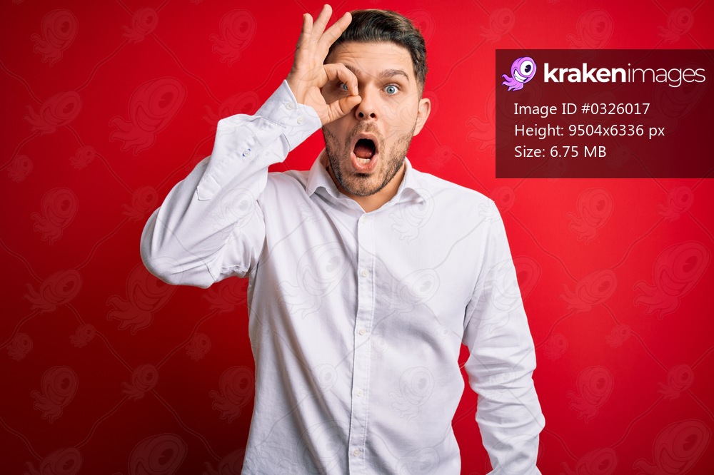 Young business man with blue eyes wearing elegant shirt standing over red isolated background doing ok gesture shocked with surprised face, eye looking through fingers. Unbelieving expression.