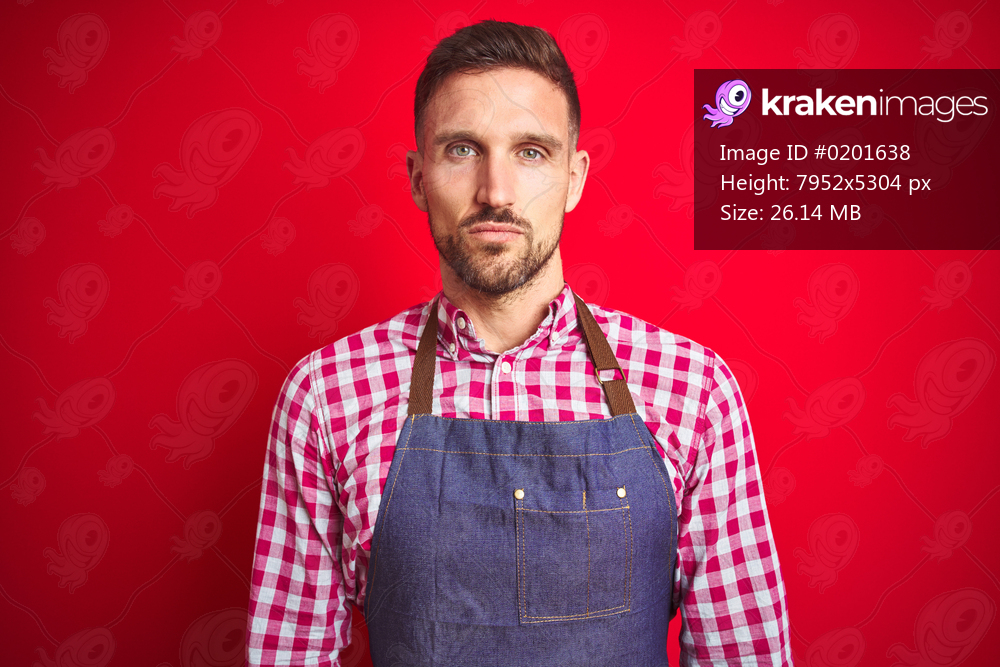 Young handsome man wearing shop owner apron uniform over red isolated background with serious expression on face. Simple and natural looking at the camera.