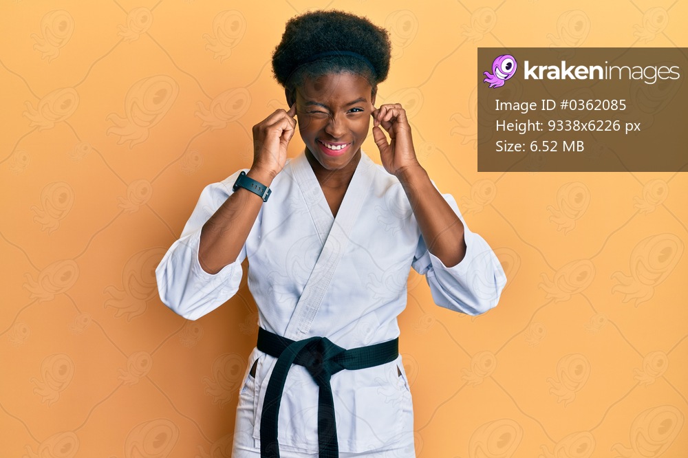 Young african american girl wearing karate kimono and black belt covering ears with fingers with annoyed expression for the noise of loud music. deaf concept. 