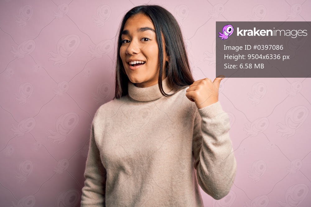 Young beautiful asian woman wearing casual turtleneck sweater over pink background smiling with happy face looking and pointing to the side with thumb up.