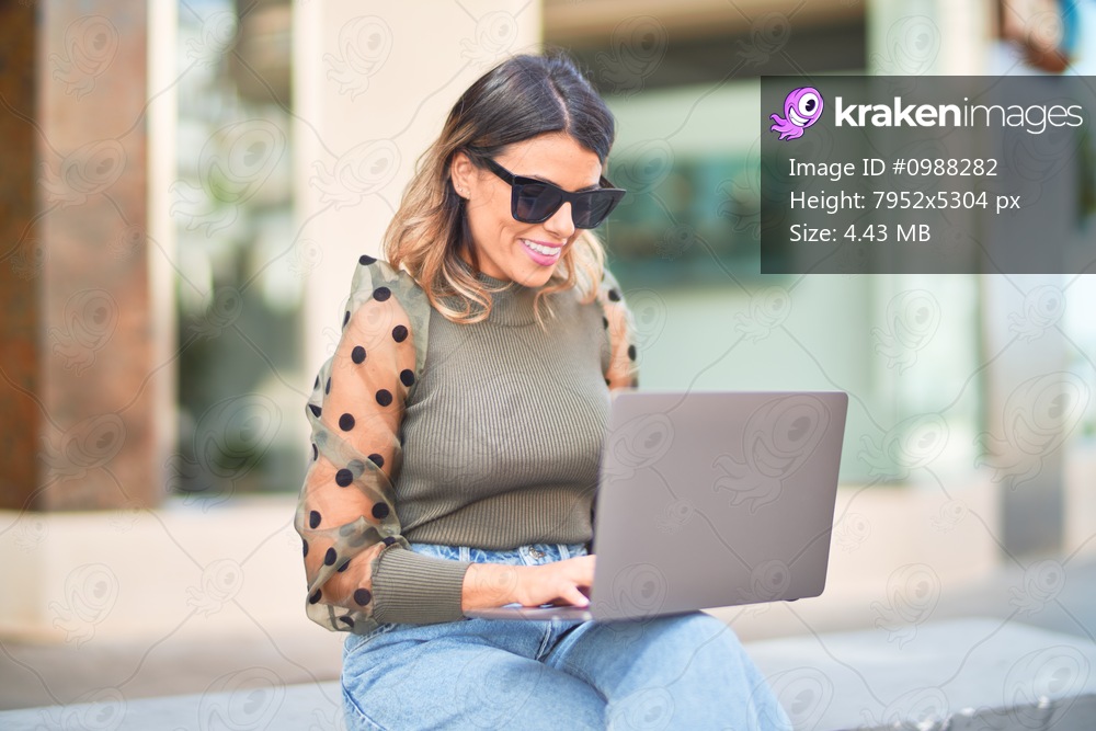 Young beautiful woman wearing sunglasses smiling happy and confident. Sitting on bench using laptop at town street