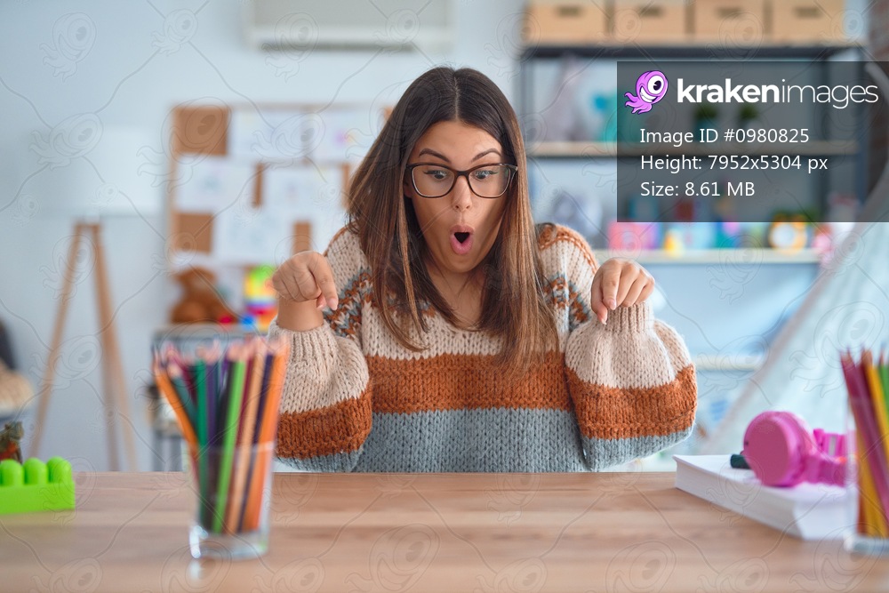 Young beautiful teacher woman wearing sweater and glasses sitting on desk at kindergarten Pointing down with fingers showing advertisement, surprised face and open mouth
