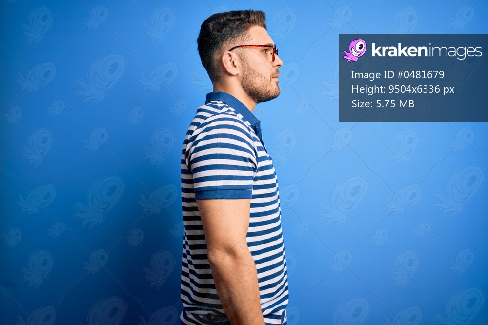Young man with blue eyes wearing glasses and casual striped t-shirt over blue background looking to side, relax profile pose with natural face with confident smile.