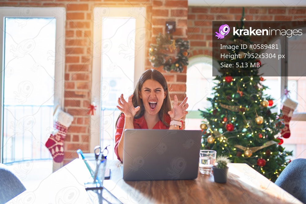 Beautiful woman sitting at the table working with laptop at home around christmas tree smiling funny doing claw gesture as cat, aggressive and sexy expression