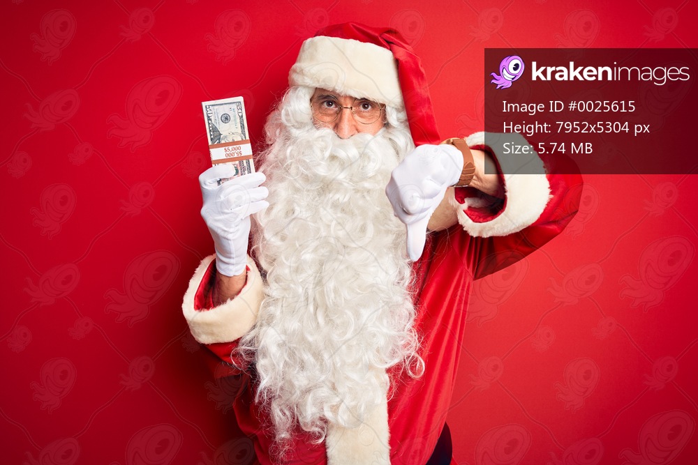 Middle age man wearing Santa Claus costume holding dollars over isolated red background with angry face, negative sign showing dislike with thumbs down, rejection concept