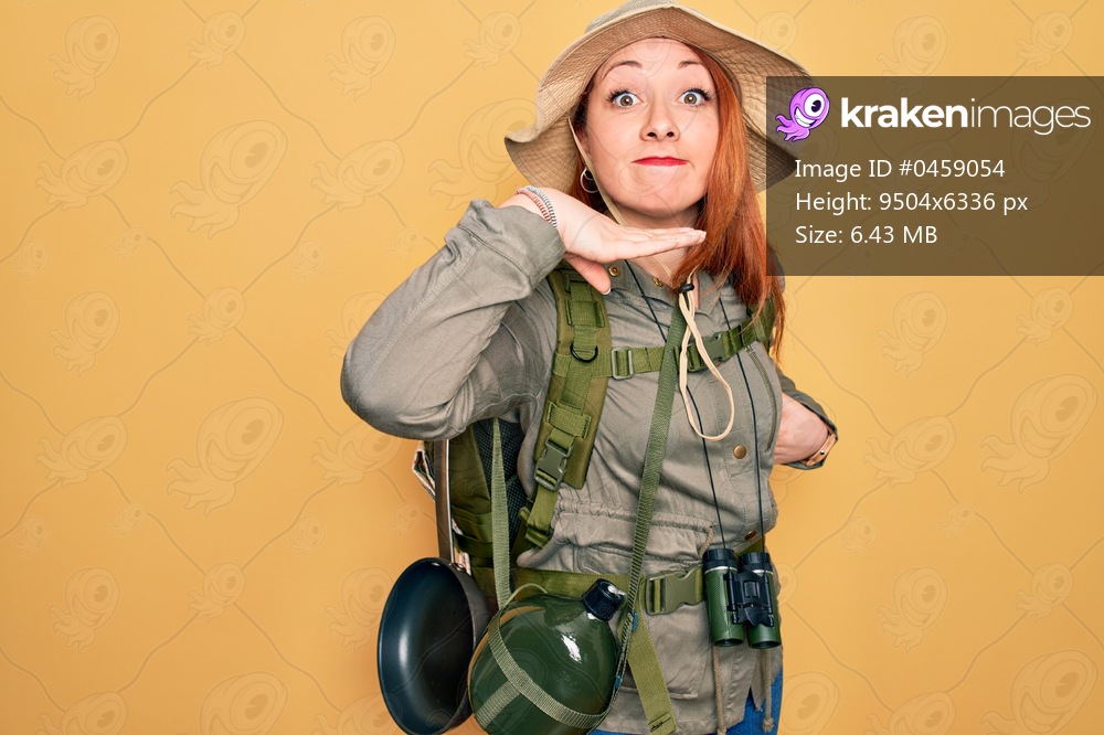 Young redhead backpacker woman hiking wearing backpack and hat over yellow background cutting throat with hand as knife, threaten aggression with furious violence