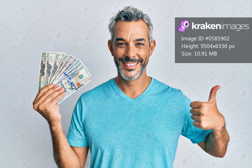 Middle age grey-haired man holding dollars smiling happy and positive, thumb up doing excellent and approval sign 