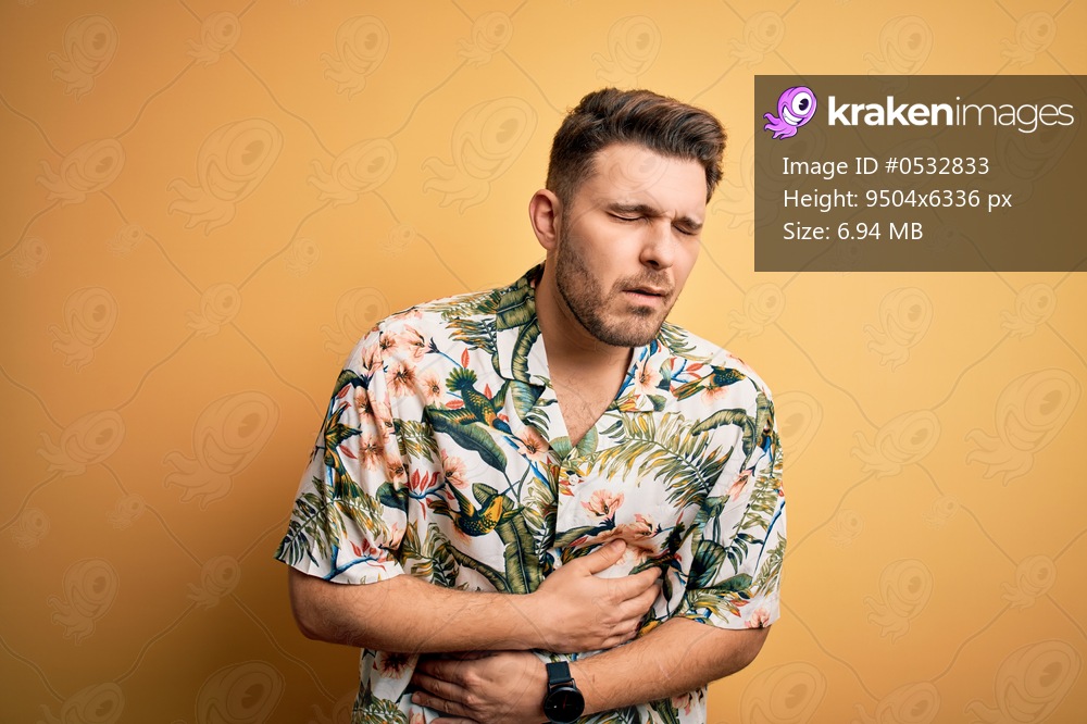 Young man with blue eyes on vacation wearing floral summer shirt over yellow background with hand on stomach because nausea, painful disease feeling unwell. Ache concept.