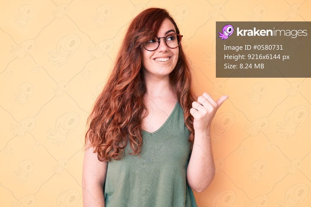 Young beautiful woman wearing casual clothes and glasses smiling with happy face looking and pointing to the side with thumb up. 