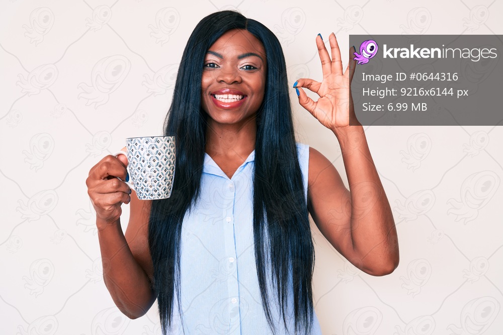 Young african american woman holding coffee doing ok sign with fingers, smiling friendly gesturing excellent symbol 
