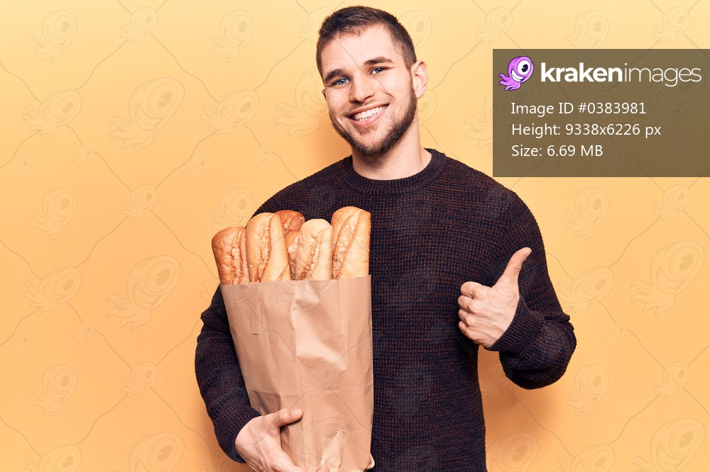 Young handsome man holding paper bag with bread smiling happy and positive, thumb up doing excellent and approval sign 