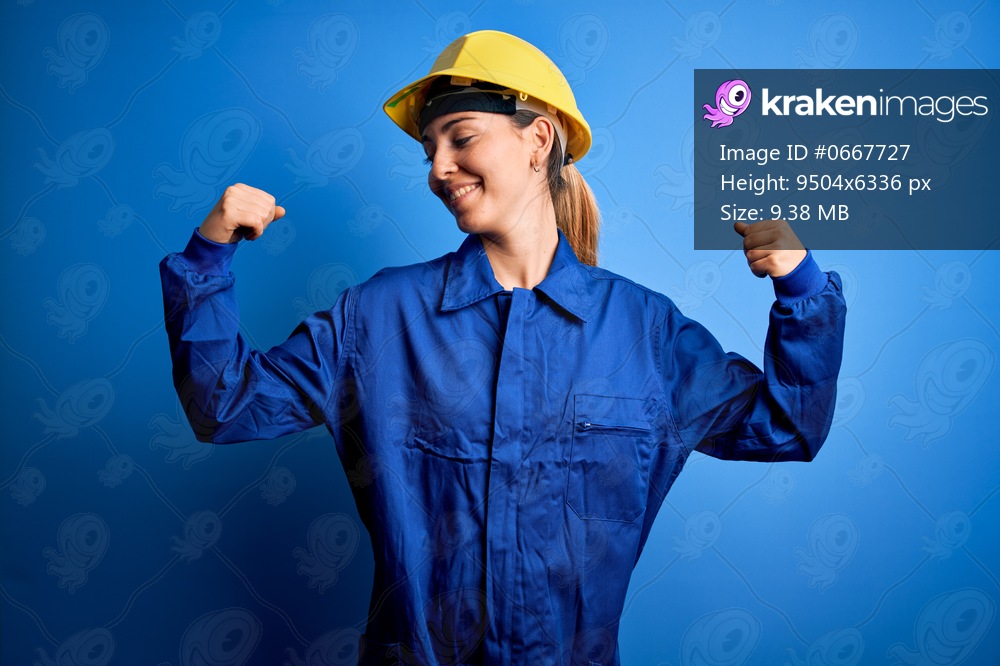 Young beautiful worker woman with blue eyes wearing security helmet and uniform showing arms muscles smiling proud. Fitness concept.