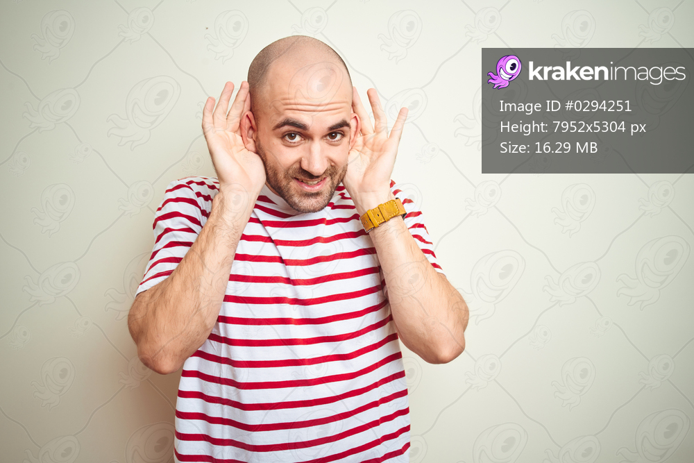 Young bald man with beard wearing casual striped red t-shirt over white isolated background Trying to hear both hands on ear gesture, curious for gossip. Hearing problem, deaf
