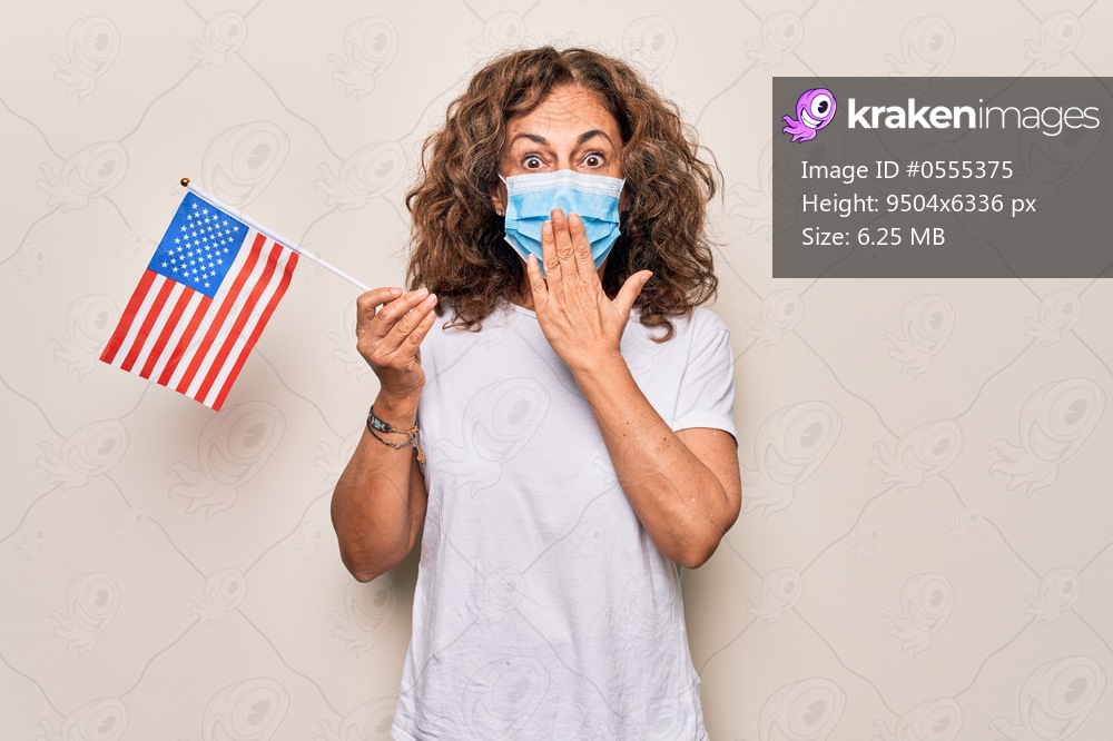 Middle age woman wearing coronavirus protection mask and holding United States of America flag covering mouth with hand, shocked and afraid for mistake. Surprised expression
