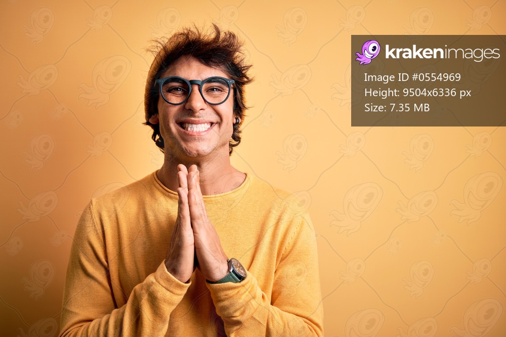 Young handsome man wearing casual t-shirt and glasses over isolated yellow background praying with hands together asking for forgiveness smiling confident.