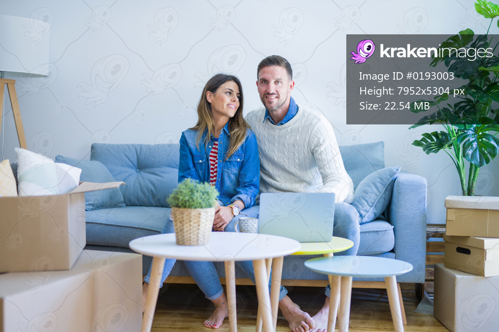Young beautiful couple sitting on the sofa drinking cup of coffee using laptop at new home around cardboard boxes