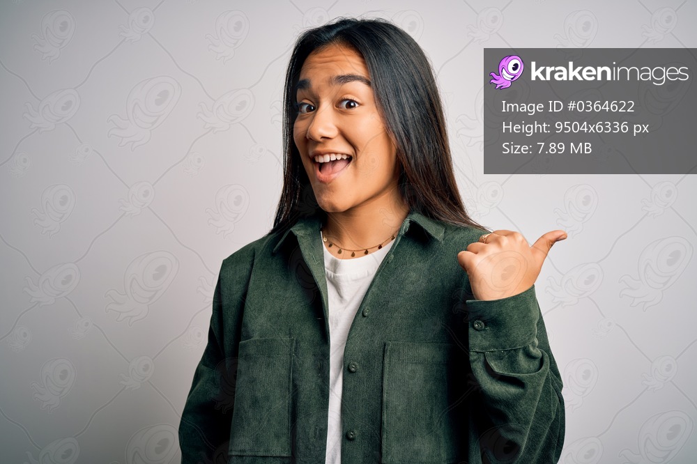 Young beautiful asian woman wearing casual shirt standing over isolated white background smiling with happy face looking and pointing to the side with thumb up.