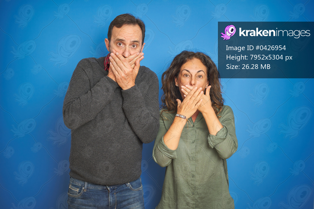 Beautiful middle age couple together standing over isolated blue background shocked covering mouth with hands for mistake. Secret concept.