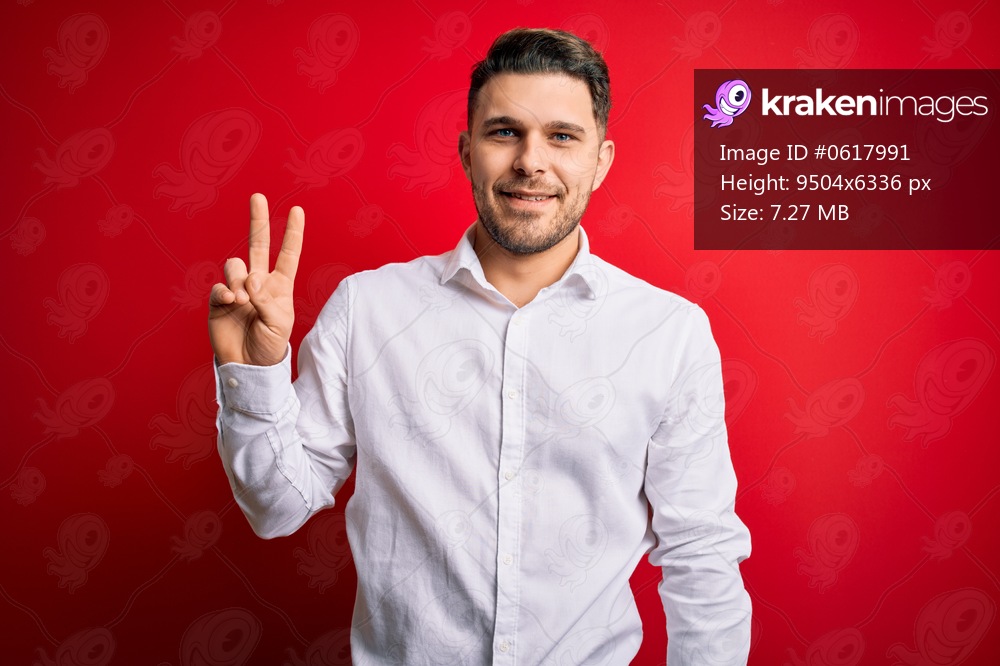 Young business man with blue eyes wearing elegant shirt standing over red isolated background smiling looking to the camera showing fingers doing victory sign. Number two.