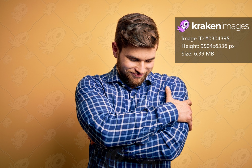 Young blond businessman with beard and blue eyes wearing shirt over yellow background Hugging oneself happy and positive, smiling confident. Self love and self care