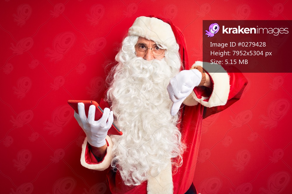 Middle age man wearing Santa Claus costume using tablet over isolated red background with angry face, negative sign showing dislike with thumbs down, rejection concept