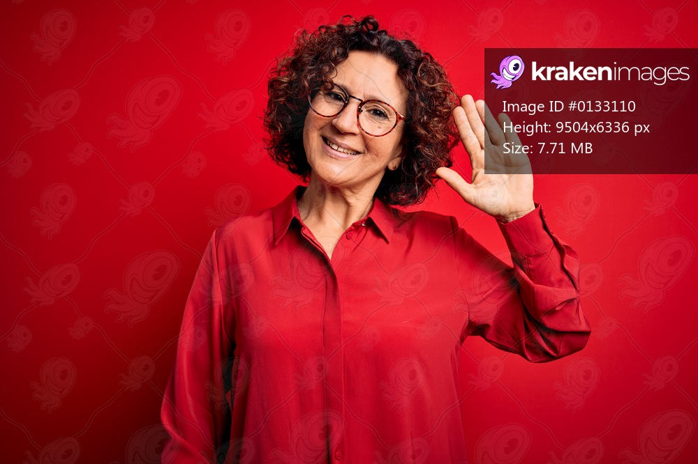 Middle age beautiful curly hair woman wearing casual shirt and glasses over red background Waiving saying hello happy and smiling, friendly welcome gesture