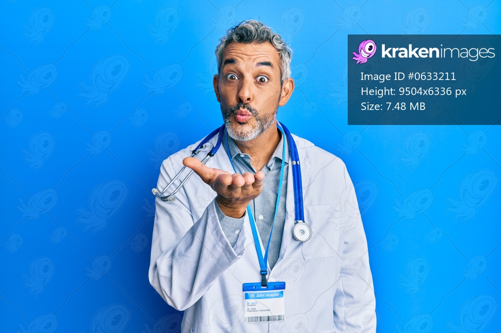 Middle age grey-haired man wearing doctor uniform and stethoscope looking at the camera blowing a kiss with hand on air being lovely and sexy. love expression. 
