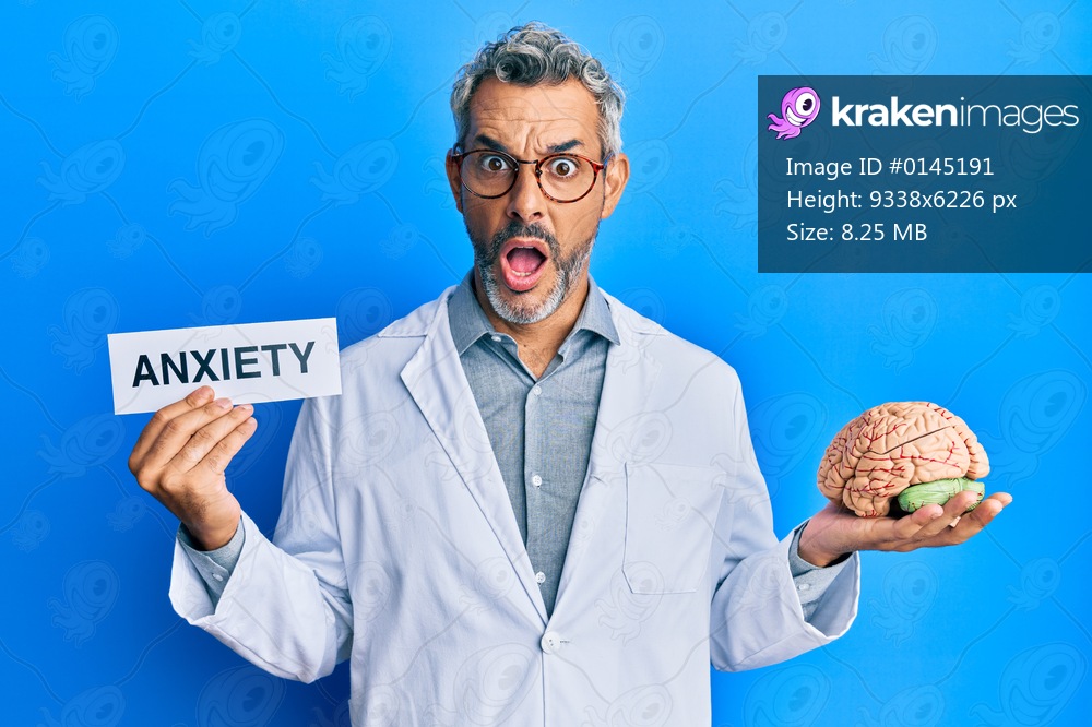 Middle age grey-haired man wearing doctor coat holding brain and anxiety message afraid and shocked with surprise and amazed expression, fear and excited face. 