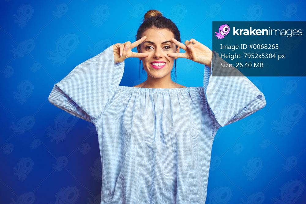 Young beautiful woman wearing bun hairstyle over blue isolated background Doing peace symbol with fingers over face, smiling cheerful showing victory