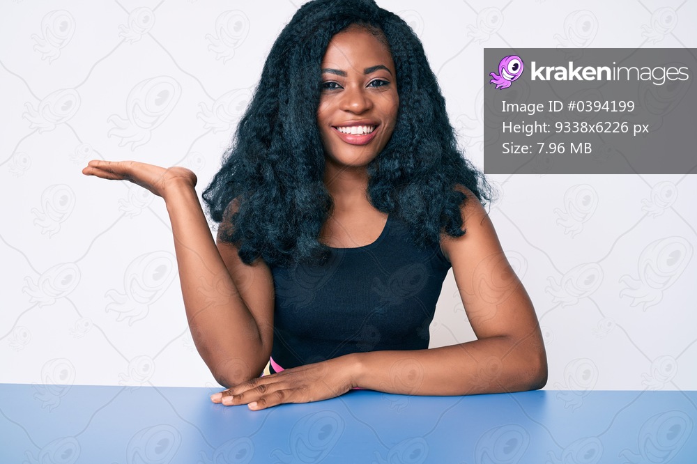 Beautiful african woman wearing casual clothes sitting on the table smiling cheerful presenting and pointing with palm of hand looking at the camera. 