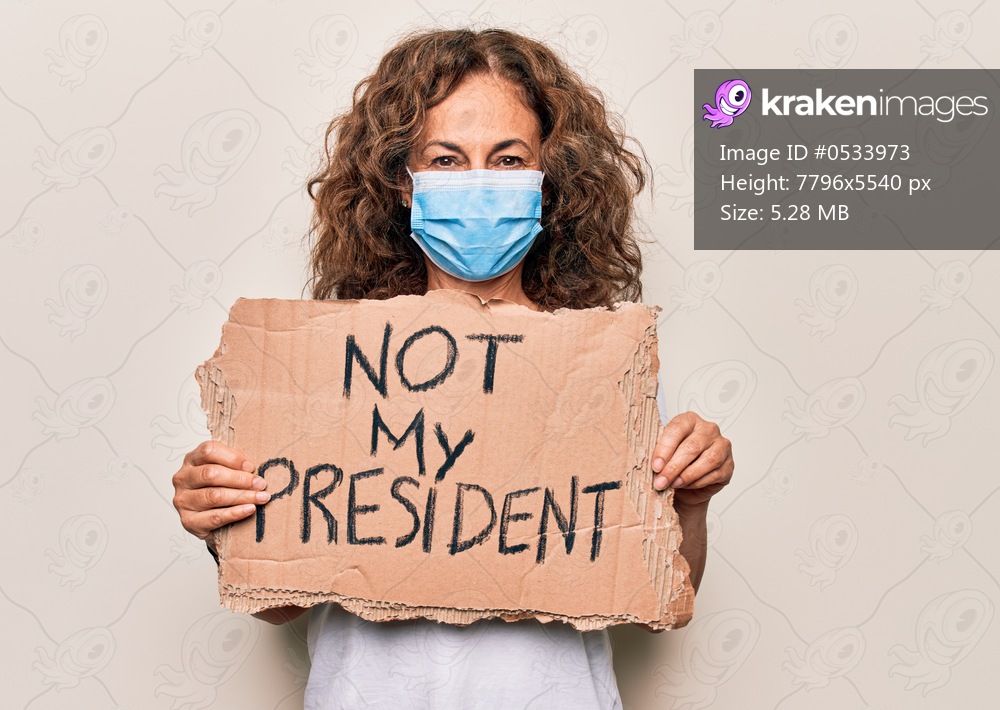 Middle age woman wearing coronavirus protection mask holding not my president protest cardboard looking positive and happy standing and smiling with a confident smile showing teeth