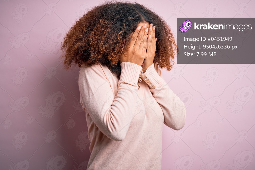Young african american woman with afro hair wearing casual sweater over pink background with sad expression covering face with hands while crying. Depression concept.