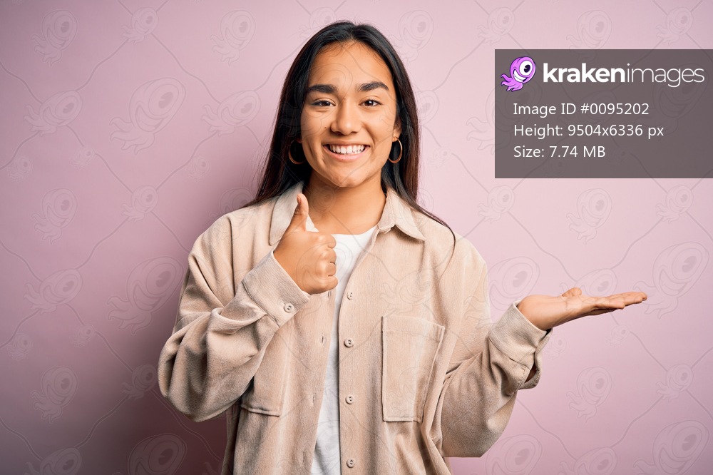 Young beautiful asian woman wearing casual shirt standing over pink background Showing palm hand and doing ok gesture with thumbs up, smiling happy and cheerful
