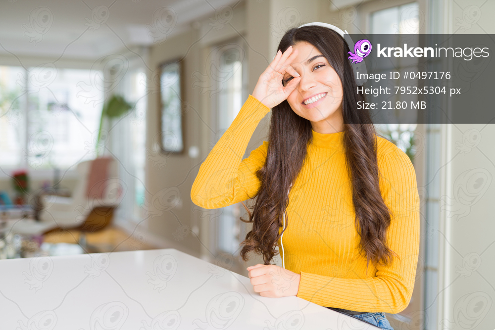 Young woman wearing headphones listening to music doing ok gesture with hand smiling, eye looking through fingers with happy face.