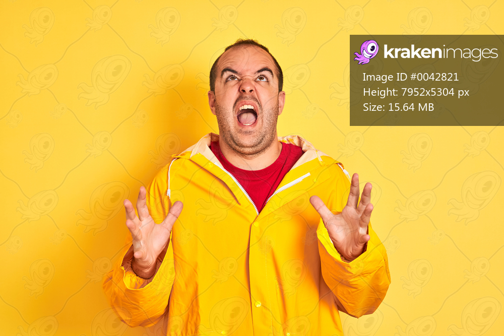 Young man wearing rain coat standing over isolated yellow background crazy and mad shouting and yelling with aggressive expression and arms raised. Frustration concept.