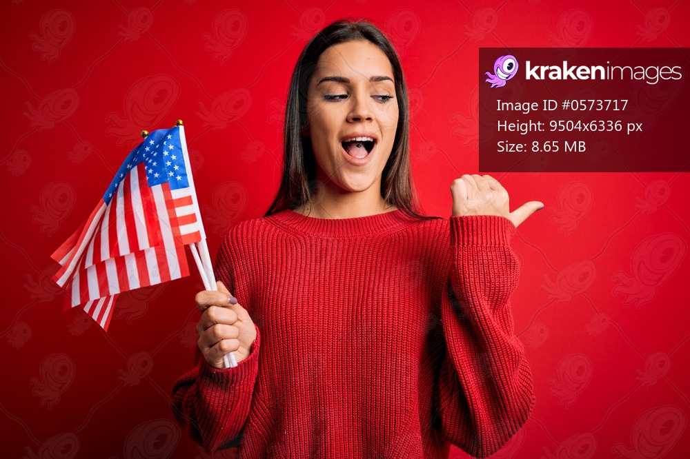Young beautiful brunette patriotic woman holding united states flag over red background pointing and showing with thumb up to the side with happy face smiling