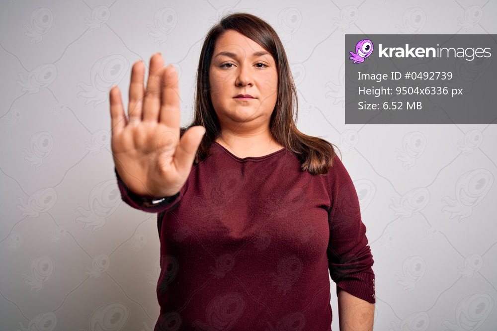 Beautiful brunette plus size woman wearing casual sweater over isolated white background doing stop sing with palm of the hand. Warning expression with negative and serious gesture on the face.