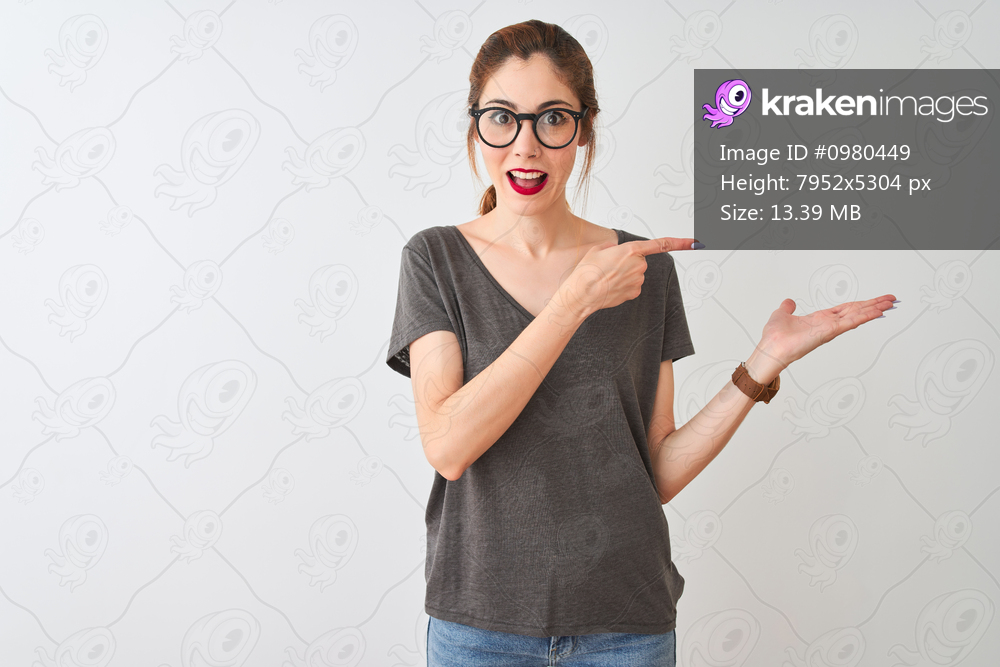 Redhead woman wearing casual t-shirt and glasses standing over isolated white background amazed and smiling to the camera while presenting with hand and pointing with finger.