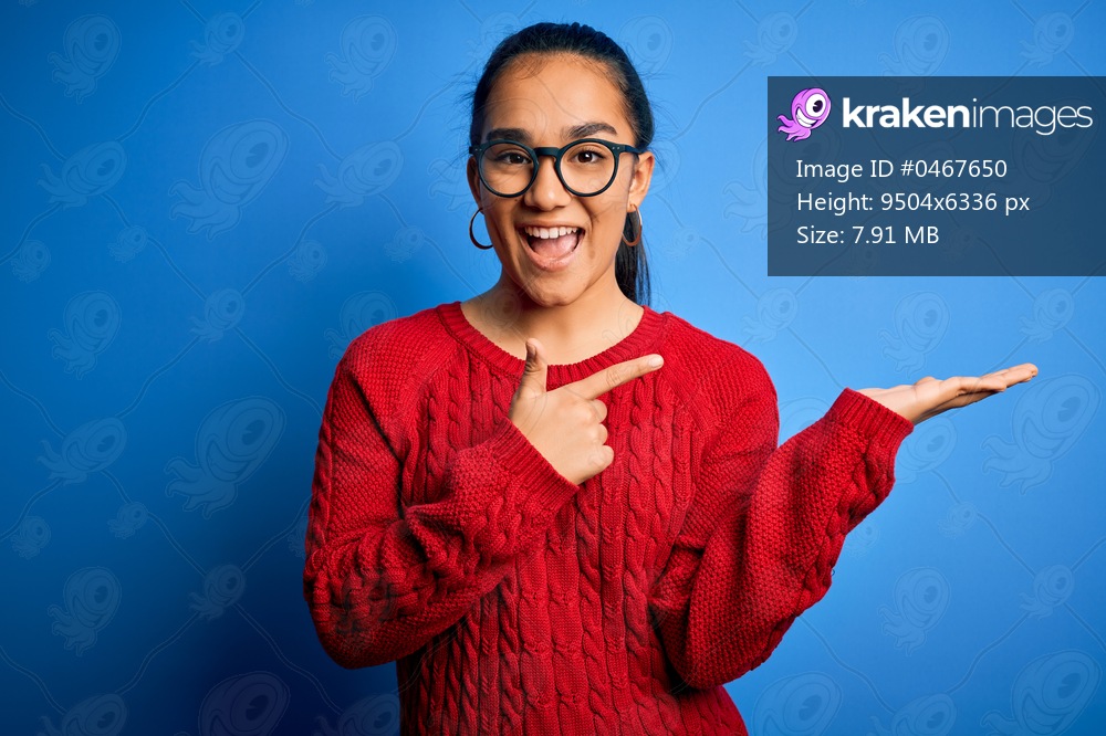 Young beautiful asian woman wearing casual sweater and glasses over blue background amazed and smiling to the camera while presenting with hand and pointing with finger.