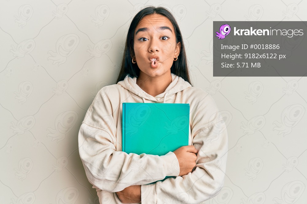 Young asian woman holding book making fish face with mouth and squinting eyes, crazy and comical. 