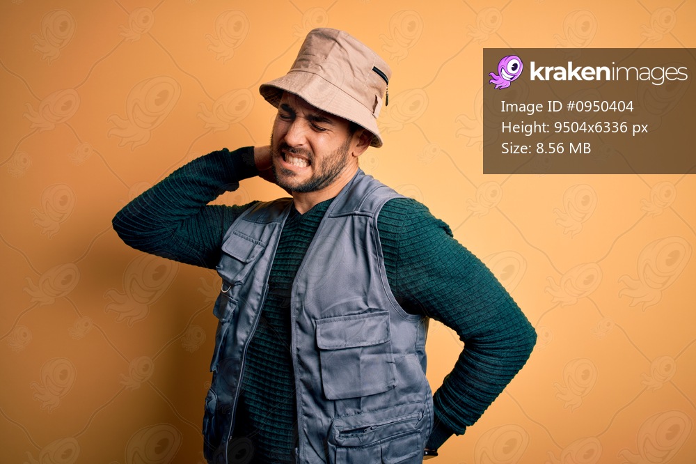 Handsome tourist man with beard on vacation wearing explorer hat over yellow background Suffering of neck ache injury, touching neck with hand, muscular pain