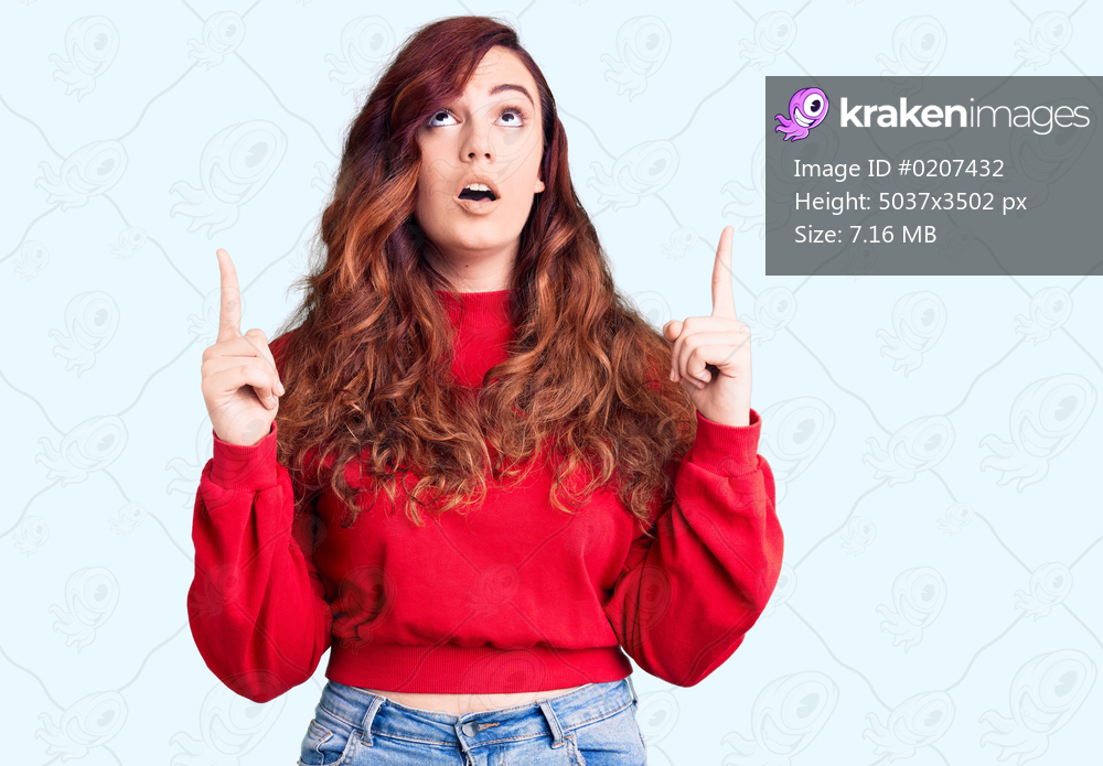 Young beautiful woman wearing casual winter sweater amazed and surprised looking up and pointing with fingers and raised arms. 