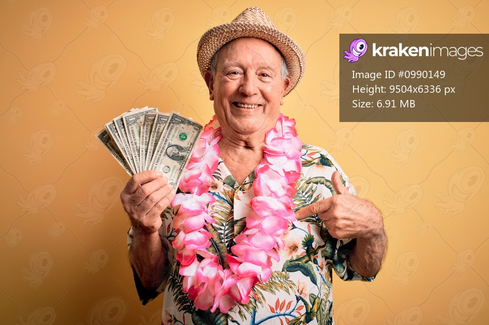 Grey haired senior man wearing summer hat and hawaiian lei holding bunch of dollars with surprise face pointing finger to himself