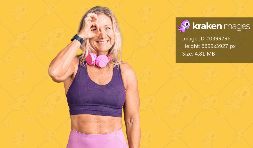 Middle age fit blonde woman wearing gym clothes and using headphones doing ok gesture with hand smiling, eye looking through fingers with happy face. 