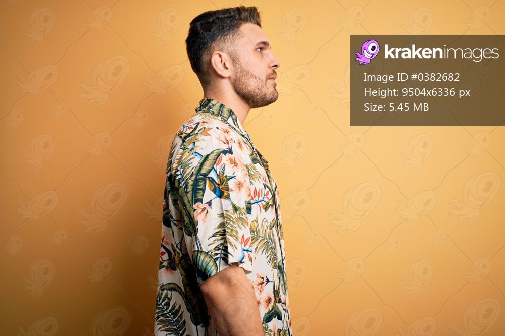 Young man with blue eyes on vacation wearing floral summer shirt over yellow background looking to side, relax profile pose with natural face with confident smile.