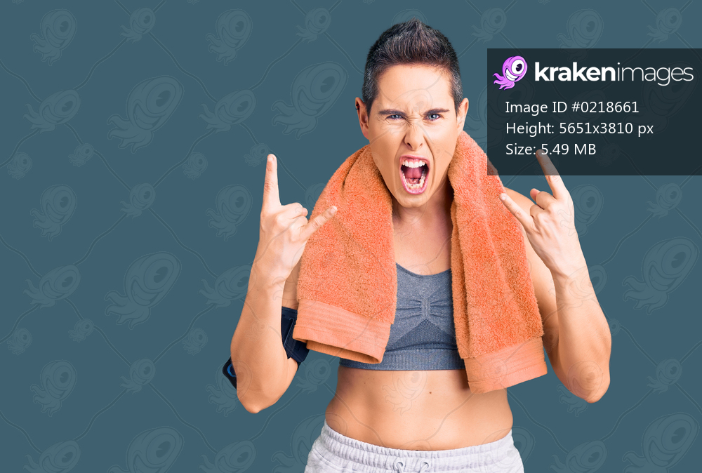 Young woman with short hair wearing sportswear and towel using smartphone shouting with crazy expression doing rock symbol with hands up. music star. heavy concept. 